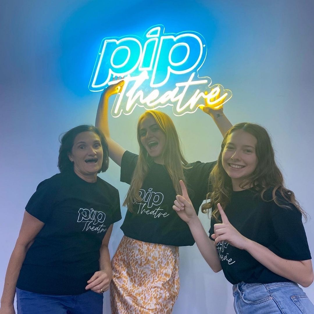 To the left is our Creative Director and founder Deidre Grace. In the middle is our Creative Developer holding a sign saying PIP Theatre above her head. To the right is the Marketing Director pointing at the sign. Everyone is smiling at the camera. Contact Us